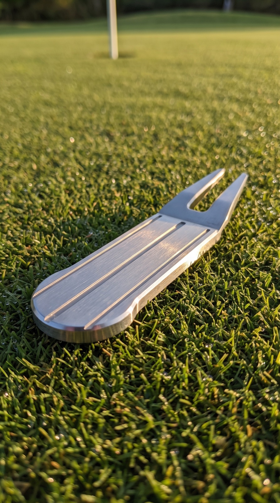 Machined stainless steel alignment tool resting on a putting green at golden hour